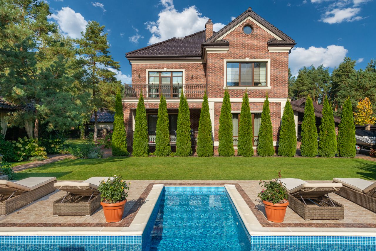 Facade of brick dwell villa with green bushes on lawn and sunbeds near swimming pool
